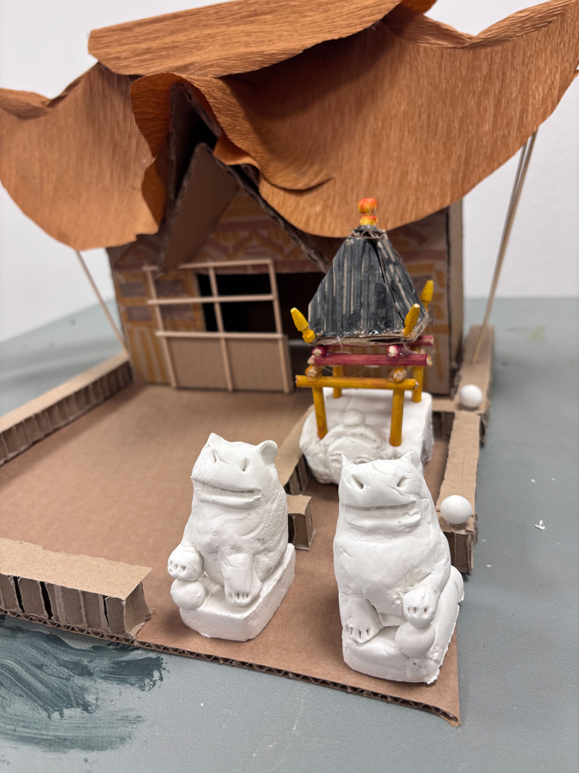 zwei Katzen aus Ton vor einem Tempel aus Papier und Pappe